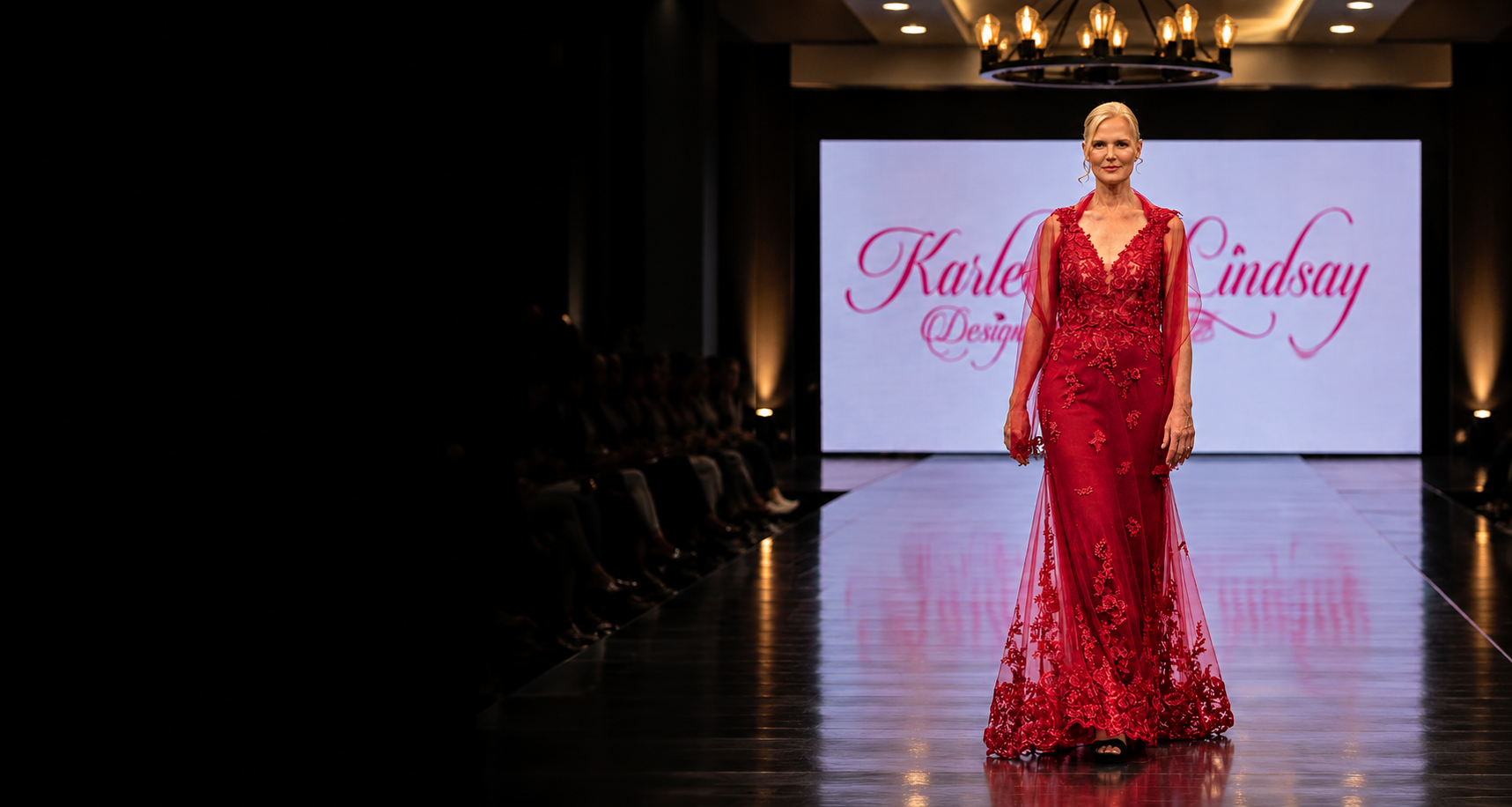 Model walks the runway in a red lace couture gown with a sheer cape.